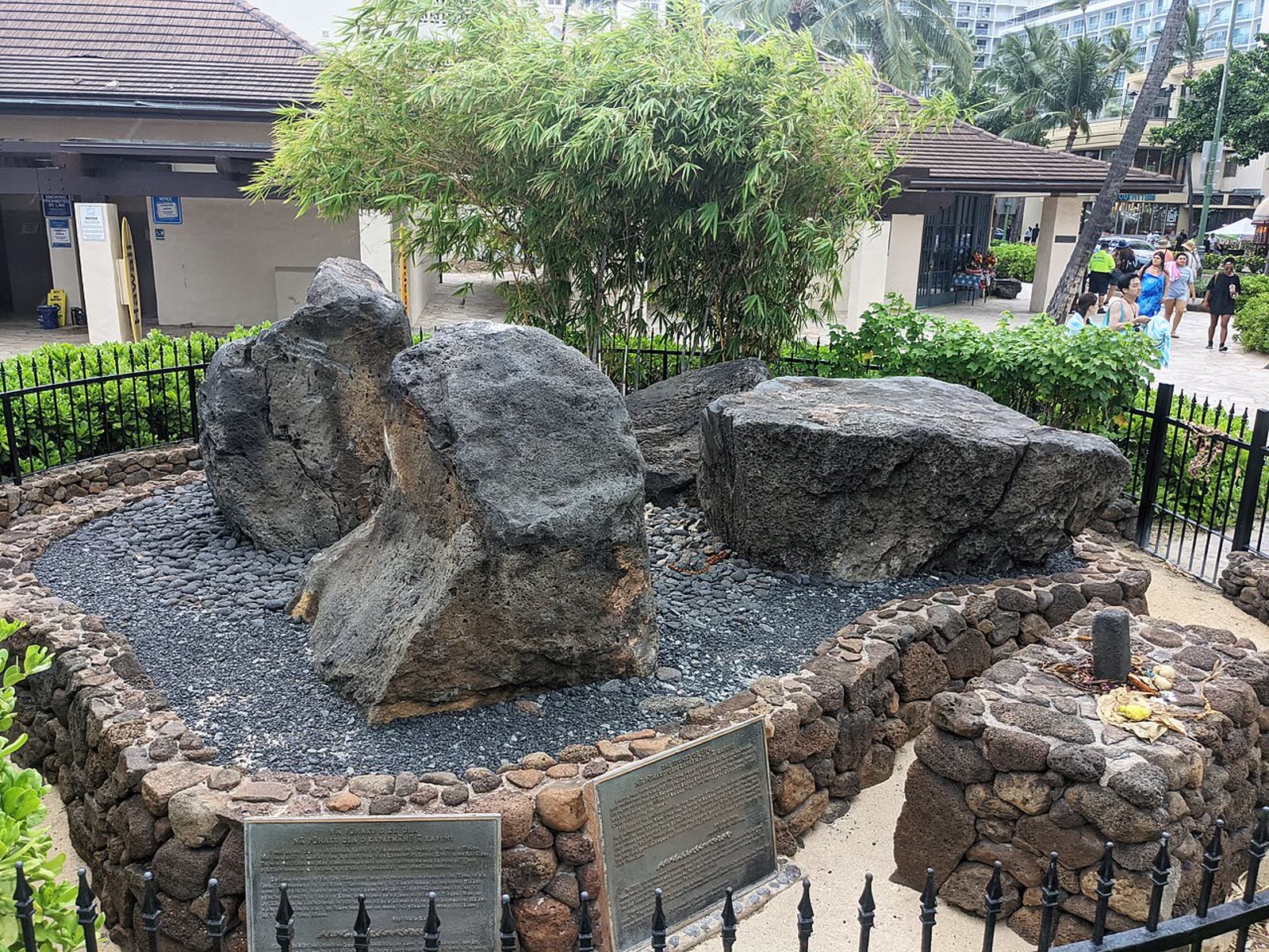 Nā Pōhaku Ola Kapaemāhū a Kapuni — four dark boulders on a stone platform behind an iron fence at Kūhiō Beach, Waikīkī